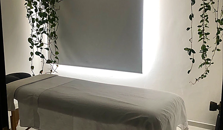 Camilla de masaje con sábanas blancas, luz de ventana y plantas colgantes
